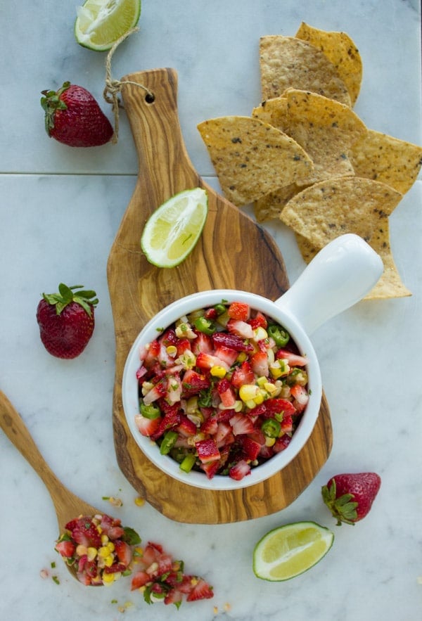 Overhead-shot of Strawberry Corn Salsa arranged on a wooden board with some chips and lime wedges