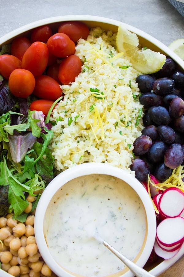 close-up of a white bowl filled with lemon cauliflower rice salad, grapes, cocktail tomatoes, chickpeas, lettuce and radish slices with a small round dish with dressing nestled in between the ingredients.