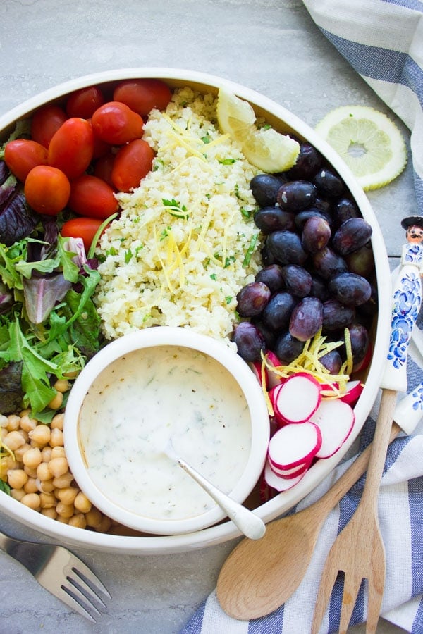 a white bowl filled with lemon cauliflower rice, grapes, cocktail tomatoes, chickpeas, lettuce and radish slices with a small round dish with dressing nestled in between the ingredients.