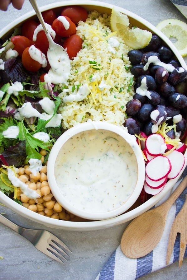 a creamy herb-speckled Yogurt Dressing being drizzled on top of a cauliflower rice salad with a small sliver spoon.