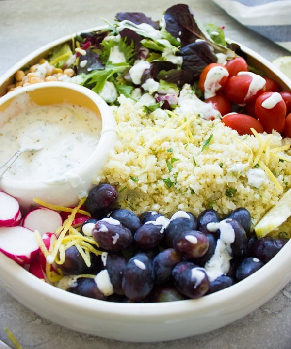 a white bowl filled with lemon cauliflower rice, grapes, cocktail tomatoes, chickpeas, lettuce and radish slices with a small round dish with dressing nestled in between the ingredients.