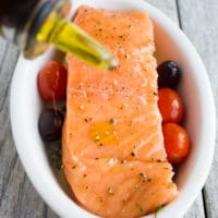 a raw salmon fillet in a small white casserole dish being drizzled with olive oil