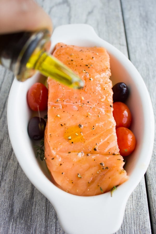 a raw salmon fillet in a small white casserole dish being drizzled with olive oil