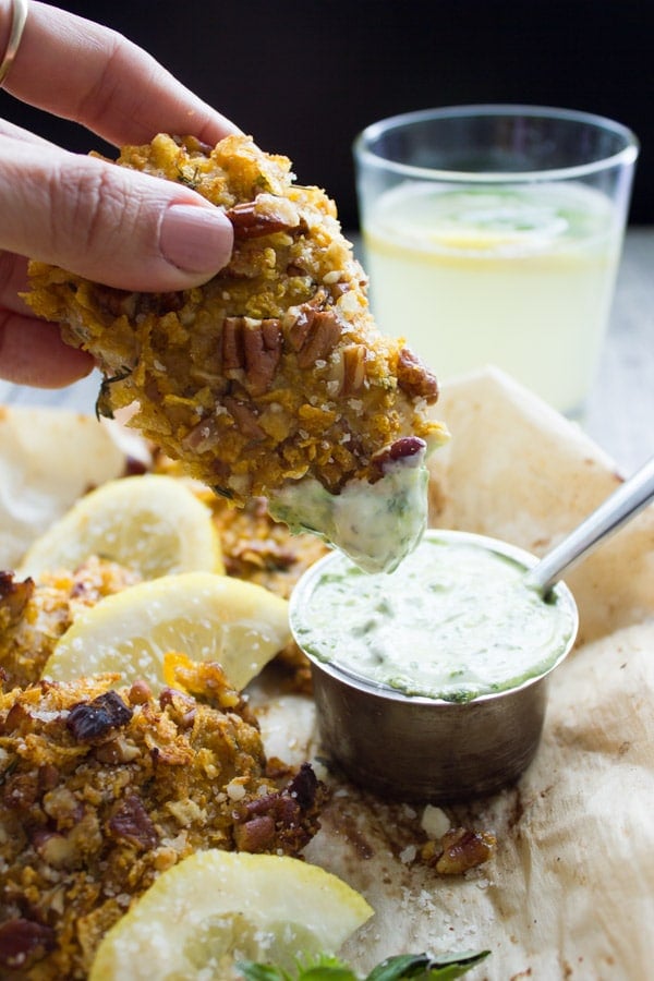 a pecan-crusted baked chicken tender being dipped into a small dish with basil dip