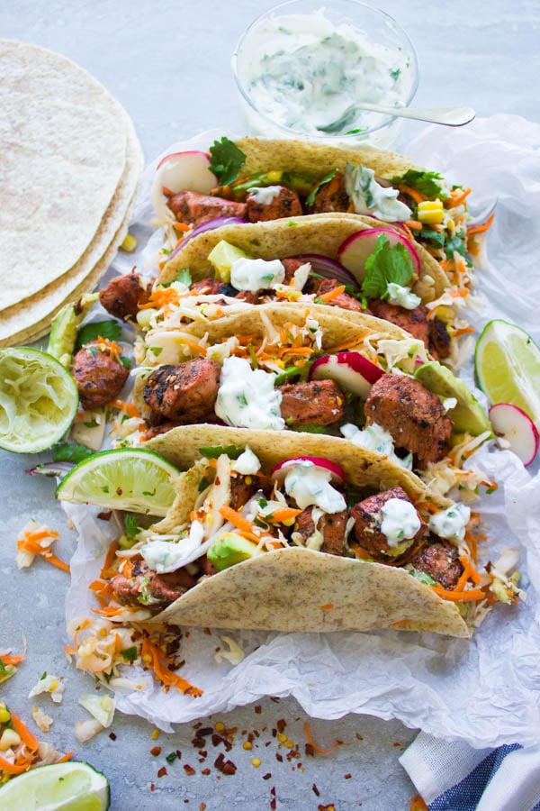 four Fiery Tandoori Chicken Tacos with Cilantro Corn Slaw and topped with yogurt dip lined up on a sheet of parchment paper
