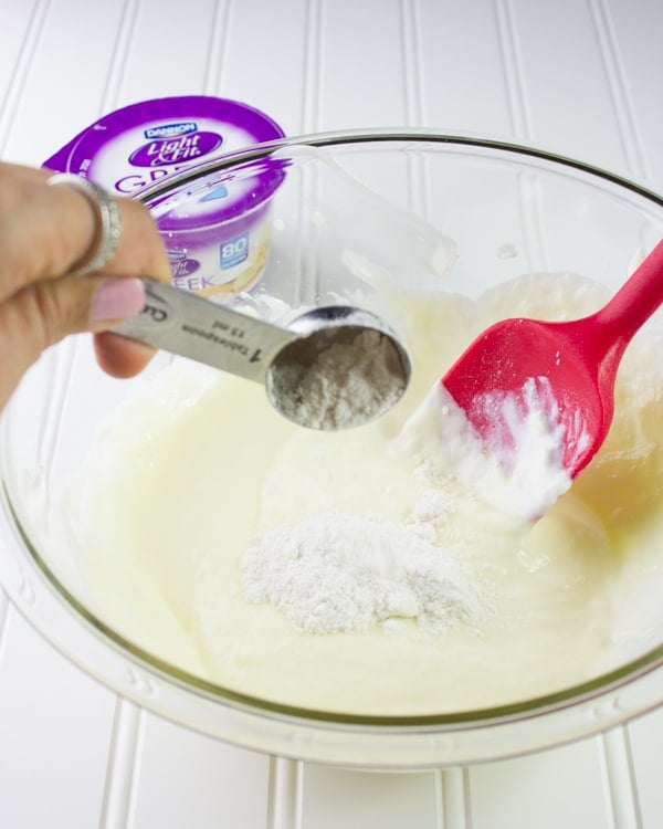 flour being added to Greek yogurt cheesecake filling in a glass bowl