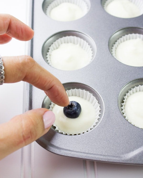 a blueberry being pushed into Greek yogurt cheesecake filling in a mini muffin tin