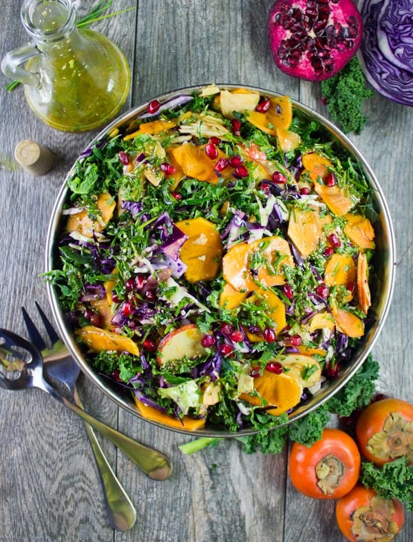 overhead shot of a big bowl of kale salad with apples, persimmon, shredded cabbage and pomegranate seeds with a dressing decanter and some salad ingredients in the background.