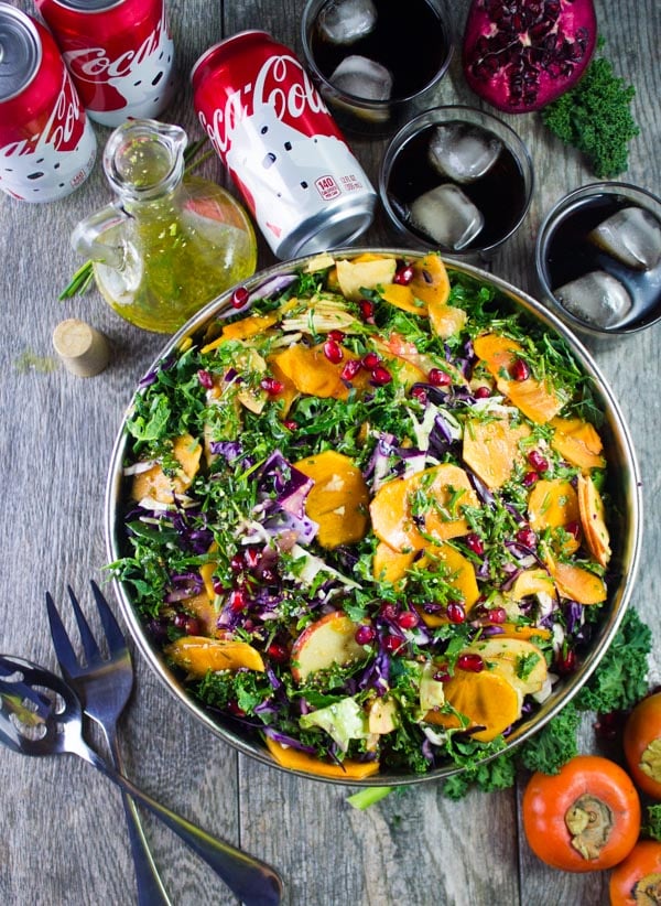 overhead shot of a big bowl of kale salad with apples, persimmon, shredded cabbage and pomegranate seeds with a dressing decanter and some salad ingredients in the background.