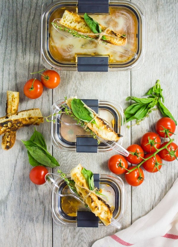 three small closed rubbermaid containers filled with lasagna soup with a breadstick on top placed on a wooden table