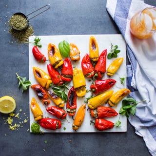 A white marble with the mini stuffed peppers on a grew board surrounded by lemons, zaatar spice, a tea towel and a punch juice