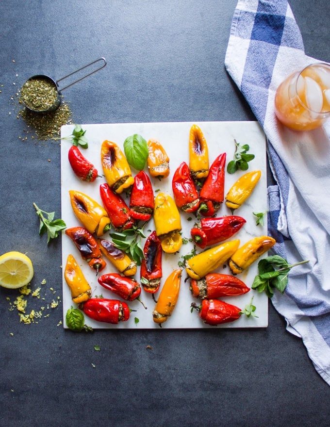 A white marble with the mini stuffed peppers on a grew board surrounded by lemons, zaatar spice, a tea towel and a punch juice