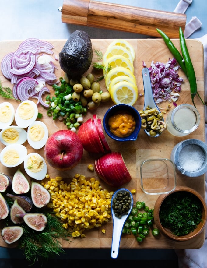 Ingredients on a wooden board for the tuna salad recipe including boiled eggs, apples, avocado, lemon juie, onions, olives, olive oil, capers, fres herbs, tomatoes, corn, tuna, jalapenos 
