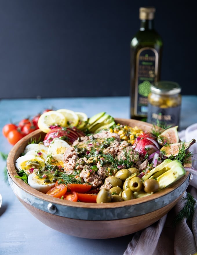 A huge bowl of tuna salad surrounded by a tea towel, a bottle of mina olive oil, some tomatoes on the board and a fork and spoon to serve