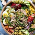 close up of a tuna salad recipe in a wooden bowl showing large chinks of tuna, boiled egs, olives, corn, avocados, tomatoes, apples, spinach and fresh herbs