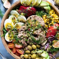 close up of a tuna salad recipe in a wooden bowl showing large chinks of tuna, boiled egs, olives, corn, avocados, tomatoes, apples, spinach and fresh herbs