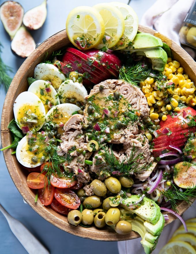 close up of a tuna salad recipe in a wooden bowl showing large chinks of tuna, boiled egs, olives, corn, avocados, tomatoes, apples, spinach and fresh herbs
