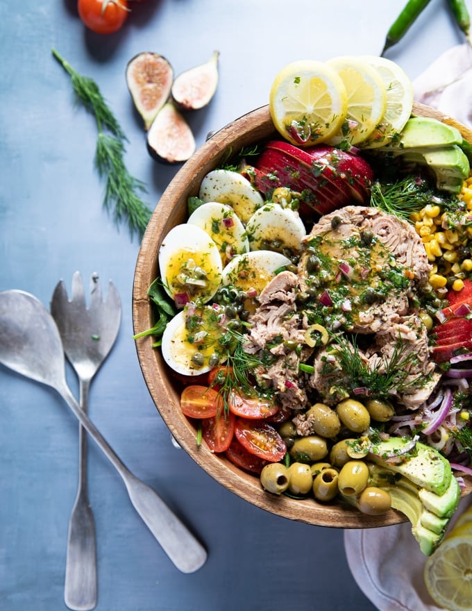 half plate of tuna salad recipe with a serving set of spoons and fork on a blue board. Fresh dill and tomatoes surrounding the big bowl of tuna salad. Salad includes chunky tuna, hard boiled eggs, apples, tomatoes, avocados, olives, lemon slices 