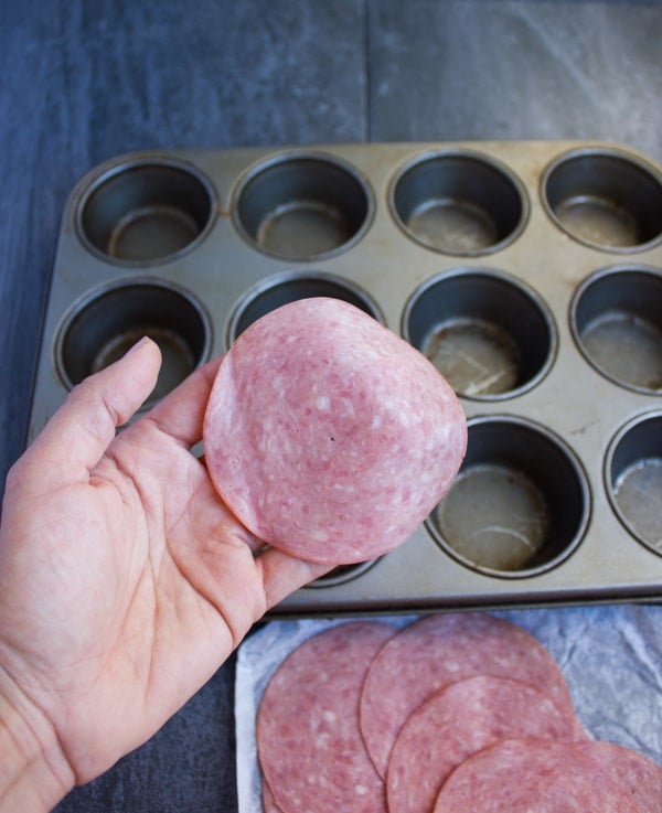 a hand holding a slice of salami with a muffin pan in the background
