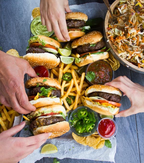 Overhead shot of 4 hands reaching for Lamb Burgers Loaded with Nacho toppings on a plate with french fries, jalapeno slices and guacamole.