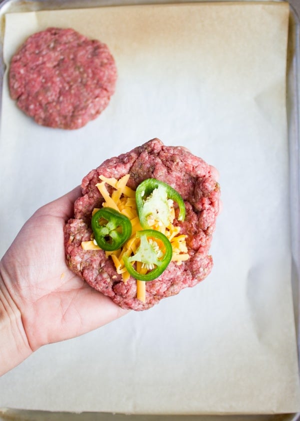 a lamb burger patty being filled with sliced jalapenos and grated cheddar cheese.