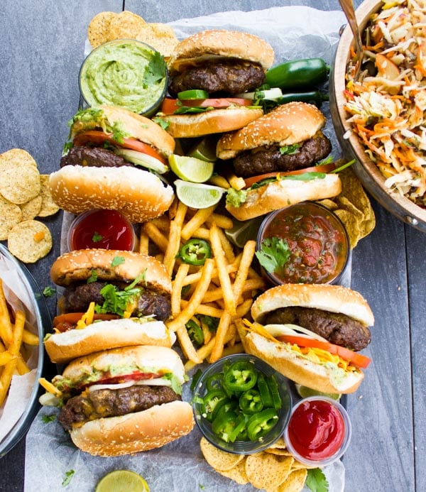 Overhead shot of Lamb Burgers Loaded with Nacho toppings on a plate with french fries and little glass bowl filled with jalapeno slices and guacamole.