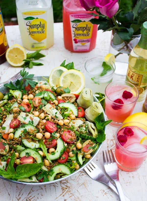 a big bowl of Mediterranean Farro Salad on a white table surrounded by bottles of lemonade and glasses with pink lemonade.