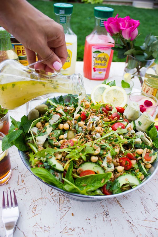 Zesty Lemonade Dressing being poured on top of a bowl of Mediterranean Farro Salad placed on a table outside.