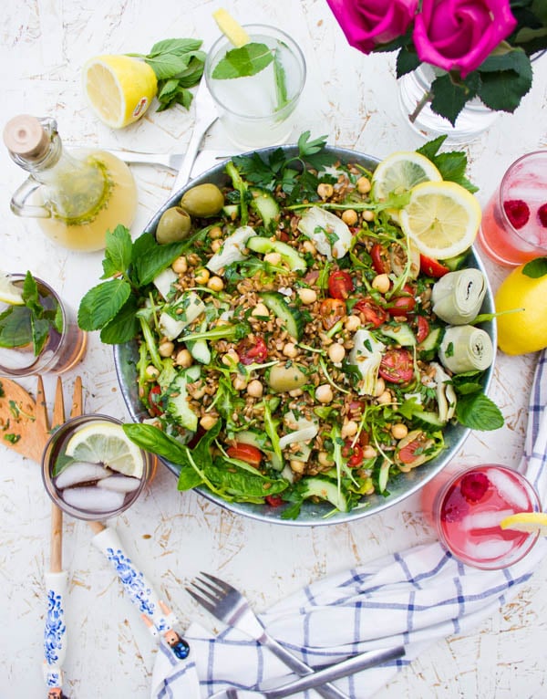 Overhead shot of a bowl of Mediterranean Farro Salad with some glasses of lemonade and wooden salad spoons surrounding it.