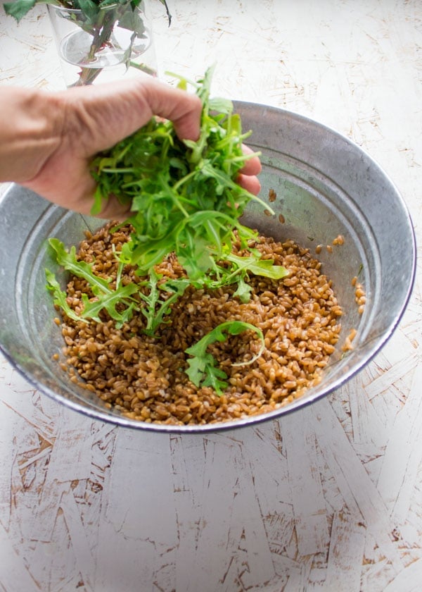 a hand full of arugula being added to a glass bowl with farro.