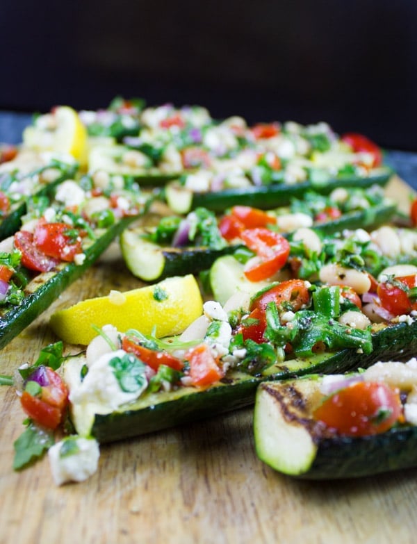 Side view of grilled Zucchini Boats loaded with White Bean Salad served on a wooden chopping board with some lemon slices on the side.