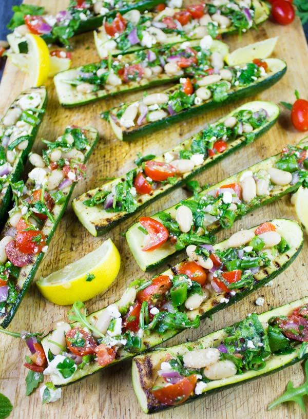 Overhead shot of grilled Zucchini Boats loaded with White Bean Salad served on a wooden chopping board with some lemon slices on the side.