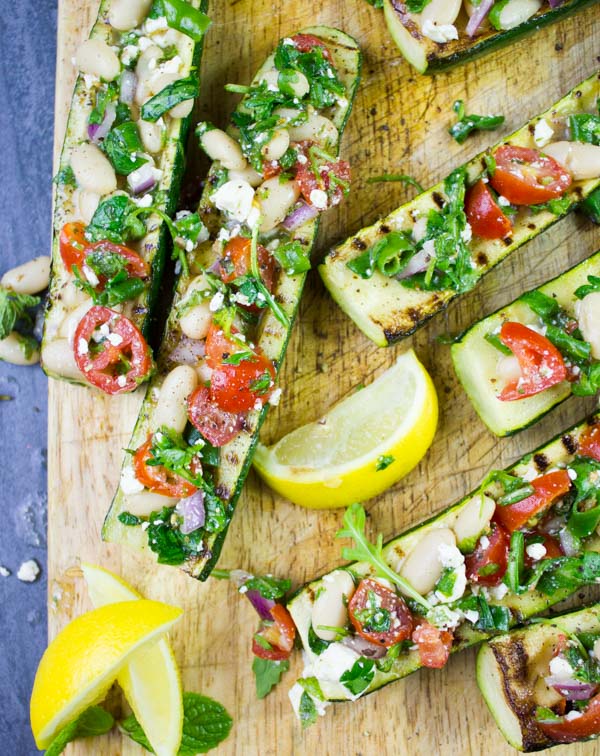 Overhead shot of grilled Zucchini Boats loaded with White Bean Salad served on a wooden chopping board with some lemon slices on the side.