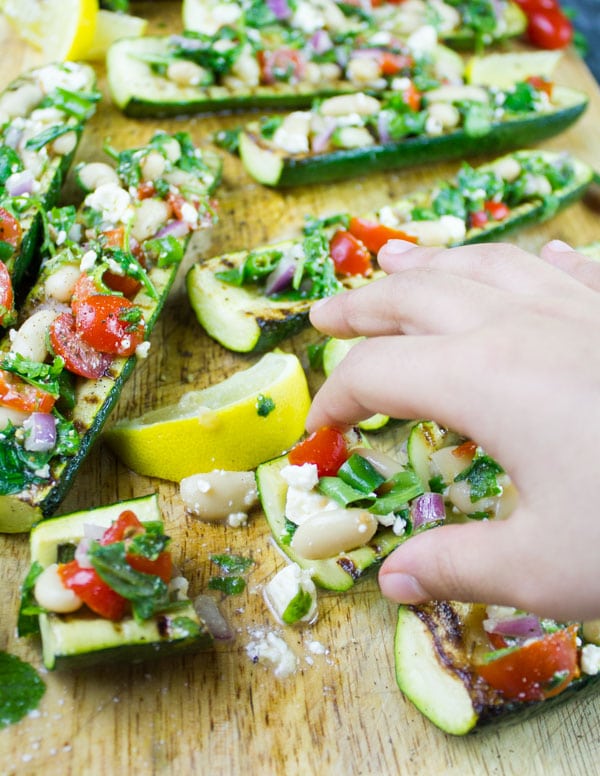 a hand reaching for a grilled Zucchini Boat loaded with White Bean Salad served on a wooden chopping board with some lemon slices on the side.