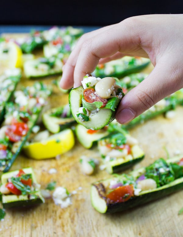 a hand holding a slice of grilled zucchini stuffed with white bean salad with more zucchini boats in the background.