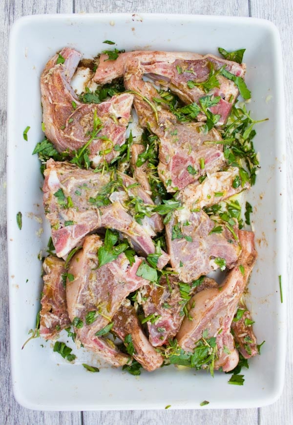 raw lamb chops marinating in a mixture of chopped herbs in a white casserole dish