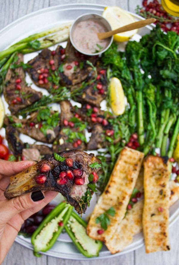 a hand holding a succulent lamb chop sprinkled with pomegranate seeds with a platter with lamb chops and grilled veggies in the background 