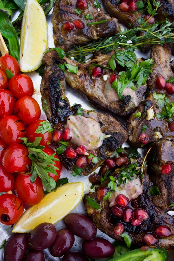 Succulent Grilled Lamb Chops with Black Olive Herb Butter sprinkled with pomegranate seeds and arranged on a silver platter with blistered cocktail tomatoes and fresh herbs