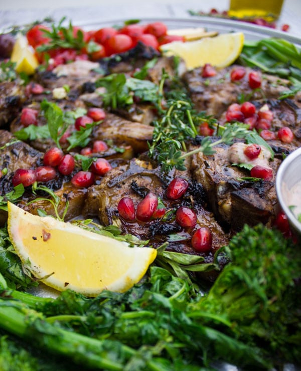 Close-up of Succulent Grilled Lamb Chops with Black Olive Herb Butter sprinkled with pomegranate seeds and arranged on a silver platter with blistered cocktail tomatoes and grilled rapini.