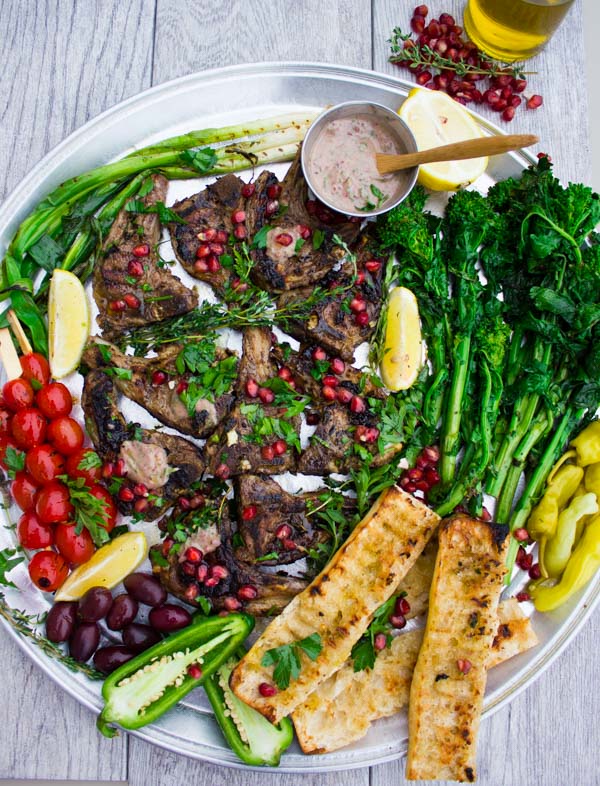 Succulent Grilled Lamb Chops with Black Olive Herb Butter sprinkled with pomegranate seeds and arranged on a silver platter with blistered cocktail tomatoes and grilled rapini as well as some charred garlic baguette.