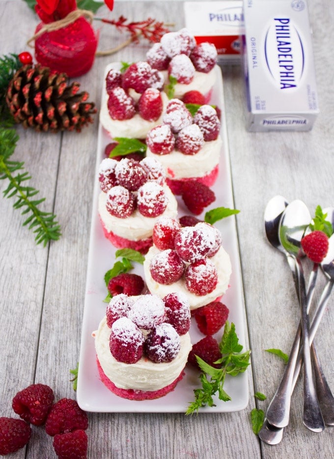 festive Mini Red Velvet Cheesecakes topped With Sugar-Dusted Raspberries on a white plate with Christmas decoration on the side