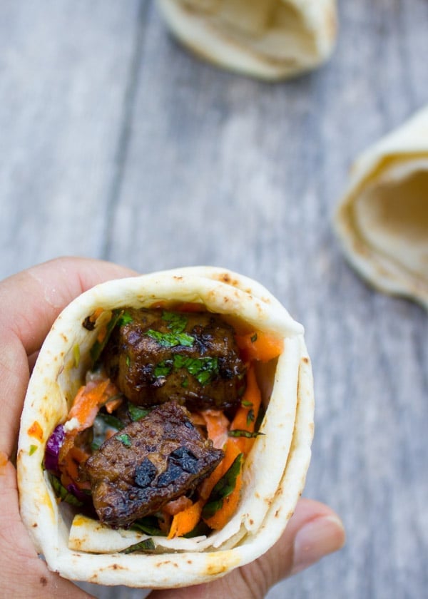 hand holding a pita cone filled with charred lamb shoulder cubes and shredded carrots.