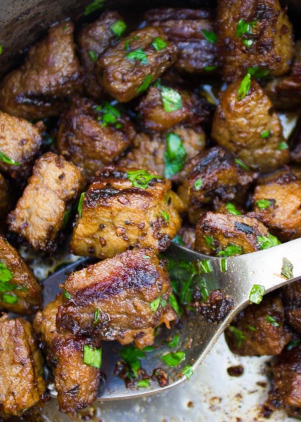 chunks of lamb shoulder being seared in a pot with chopped herbs.