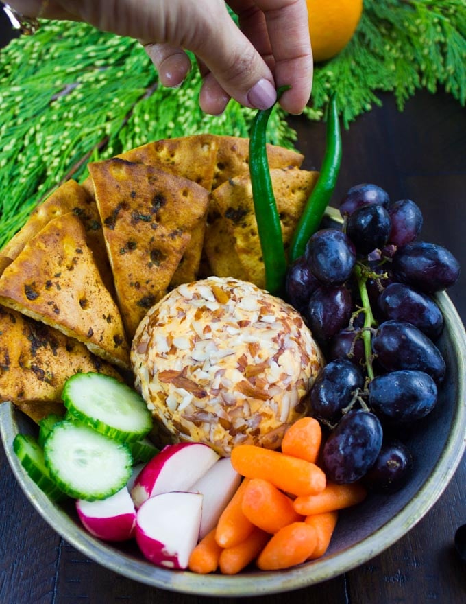 a nut-crusted cheddar cheese ball in a bowl surrounded with fruit, homemade pita chips and veggies