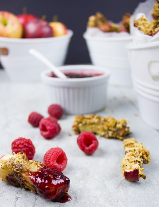 granola-coated baked apple fries lying on a kitchen counter with the front fry dipped in raspberry sauce, more fries and fresh raspberries and apples in the background