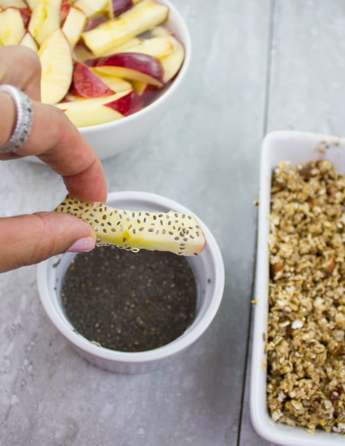 an apple stick being dipped into chia seeds before coating it with granola 