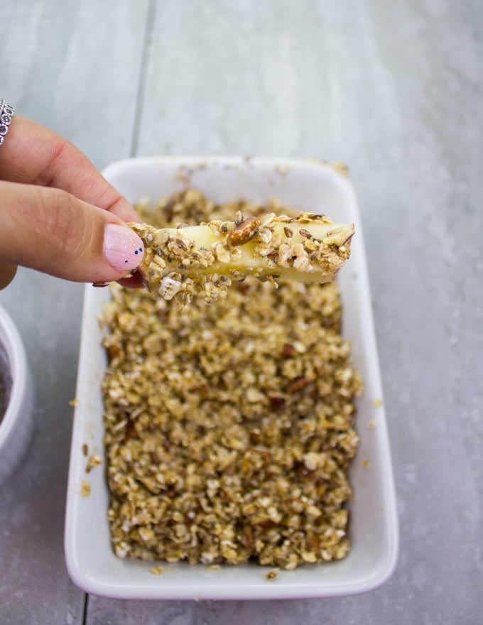 an apple stick being rolled around in a dish with granola before baking it into apple fries
