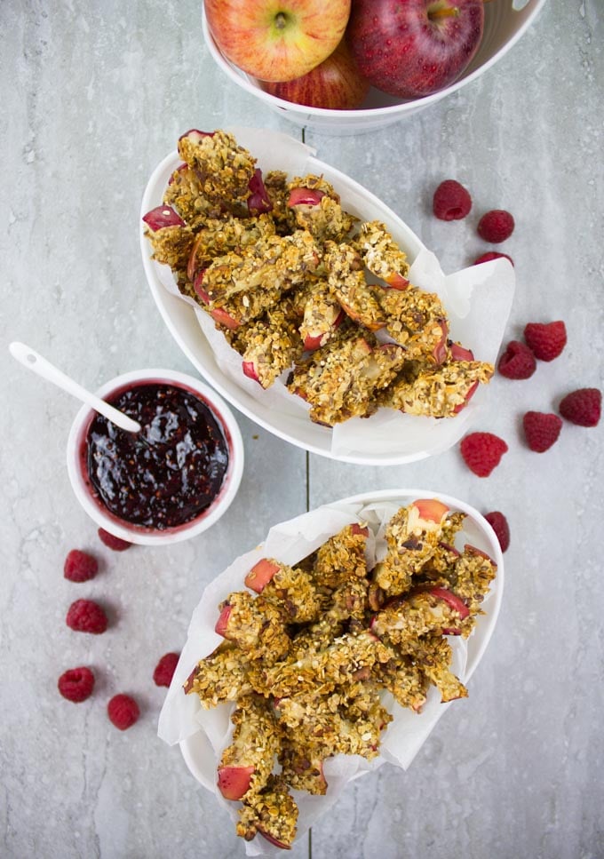 overhead shot of two oval dishes filled with baked apple fries next to some fresh raspberries, a bowl with apples and a side dish with raspberry sauce