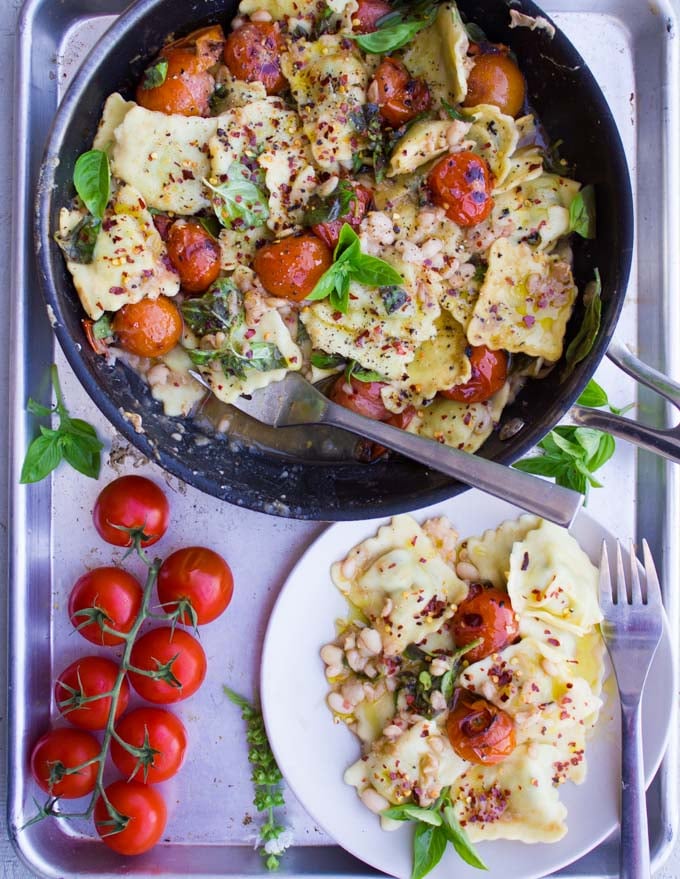 a serving of 15 Minute One Pan Ravioli with navy beans and mini tomatoes sprinkled with chili flakes and fresh basil on a white plate in front of a skillet with more ravioli