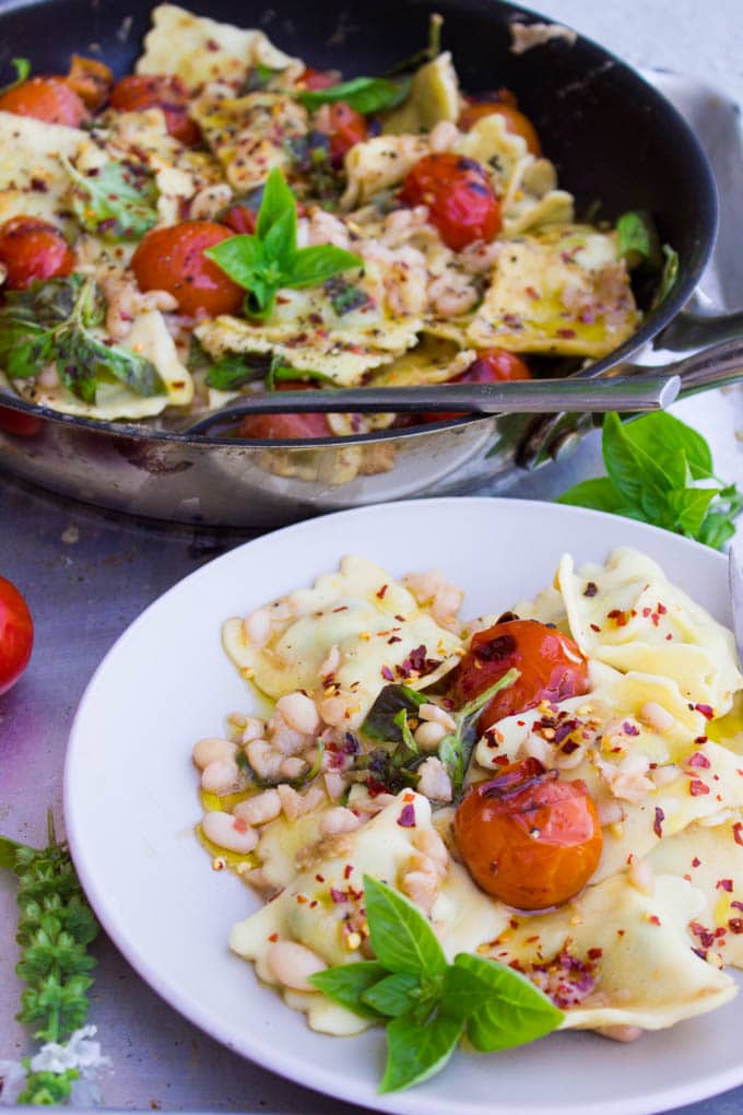 a serving of 15 Minute One Pan Ravioli with navy beans and mini tomatoes sprinkled with chili flakes and fresh basil on a white plate in front of a skillet with more ravioli